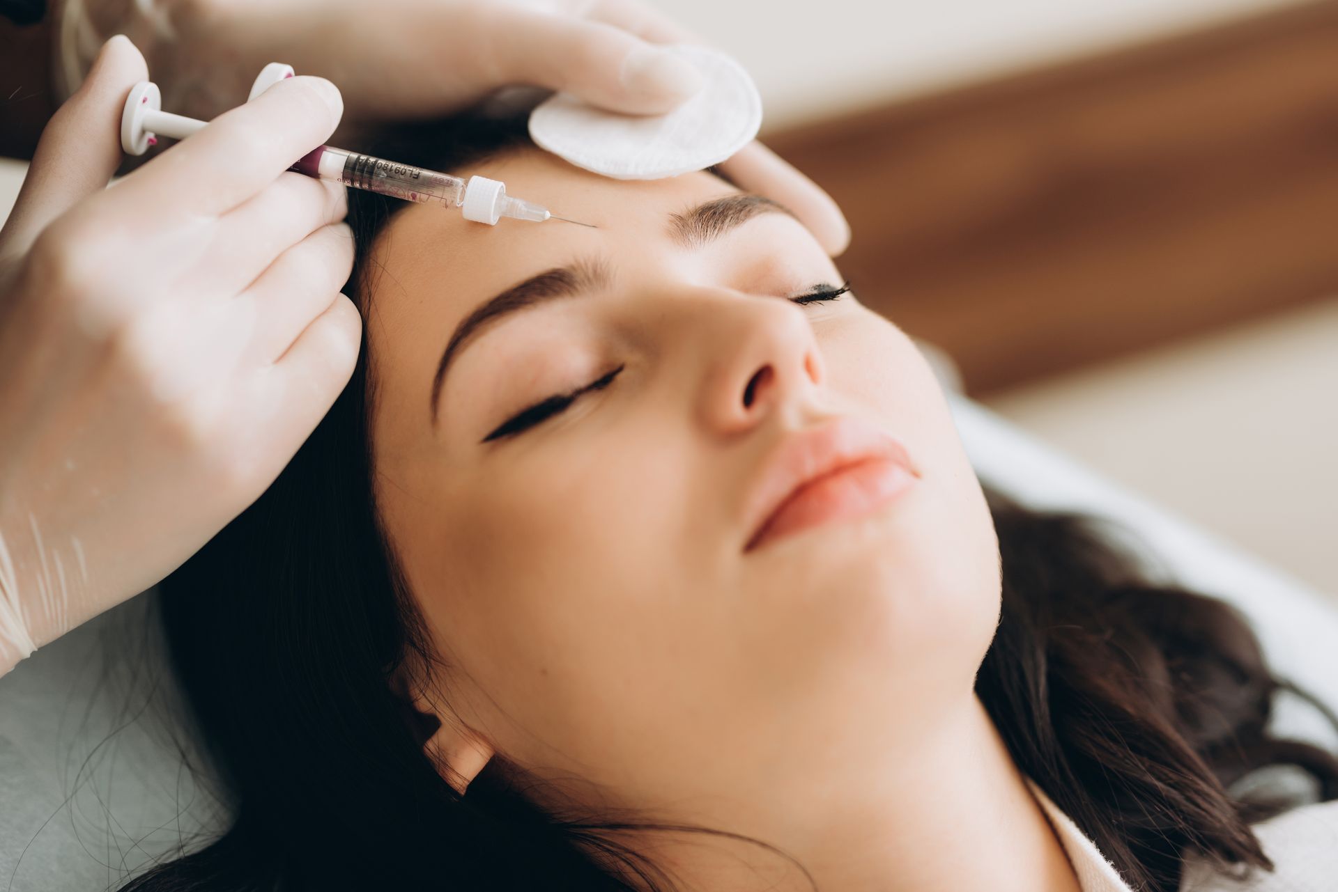 Woman receiving a cosmetic injection in her forehead; medical setting, closed eyes.