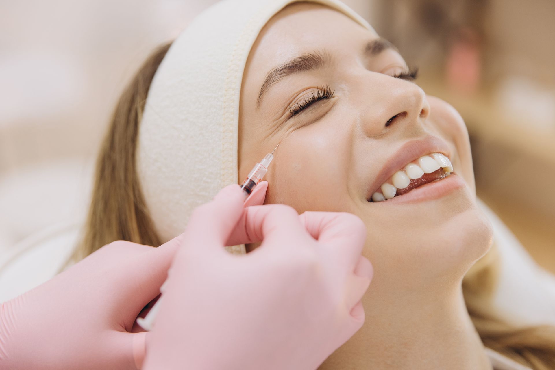 A person receiving a facial injection. A gloved hand injects near their eye. They smile with eyes closed, indoors.