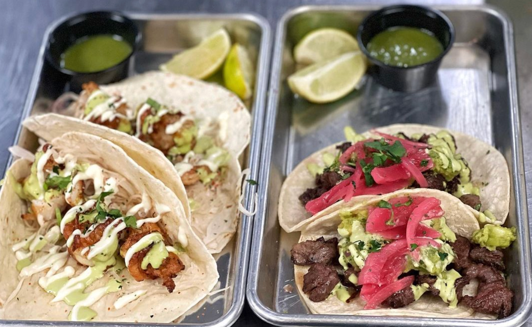 Two metal trays filled with tacos and sauces on a table.