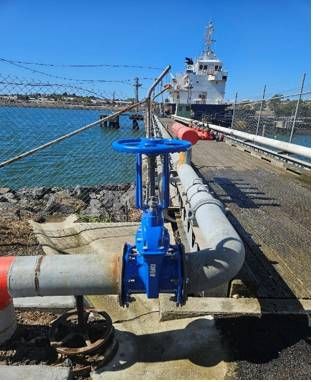 A blue valve is sitting on top of a pipe next to a body of water.