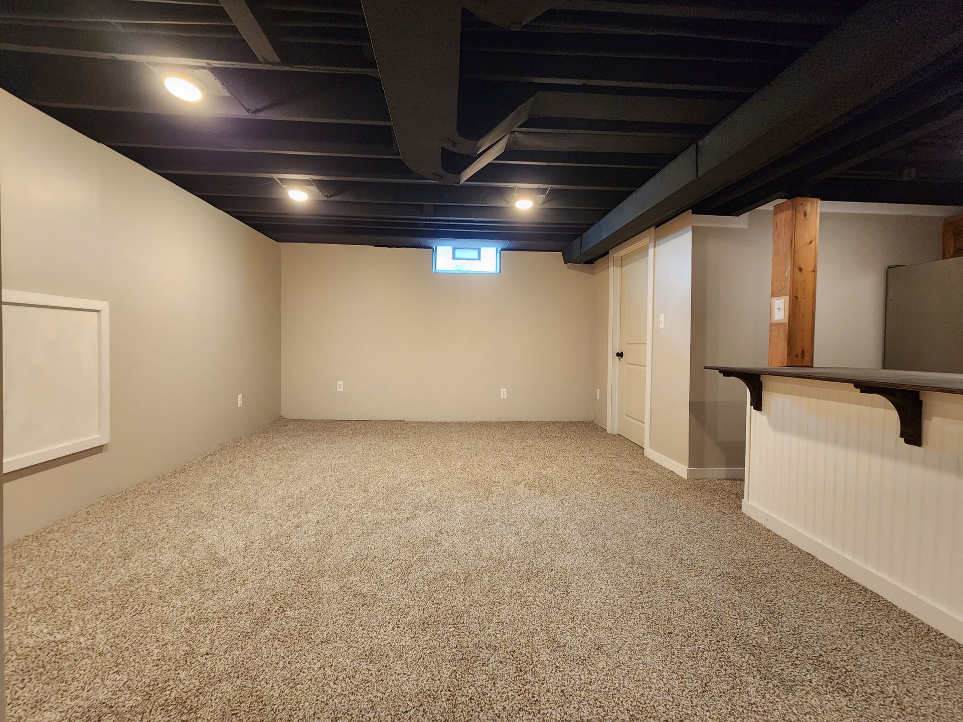 Empty basement room with beige carpet, white walls, exposed black ceiling beams, and a small window.