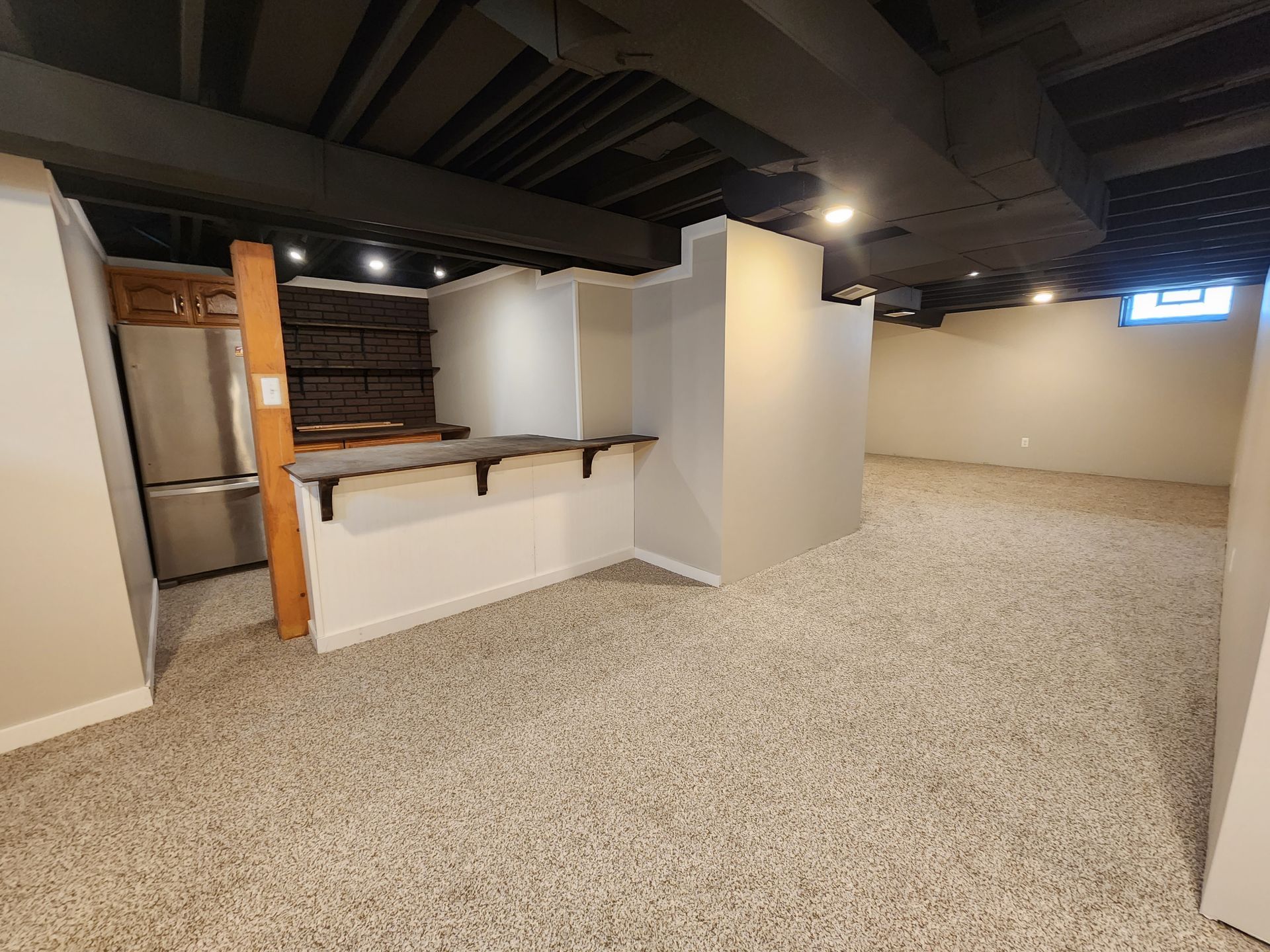 Empty basement room with carpet, exposed ceiling beams, and a small white bar area with shelves.