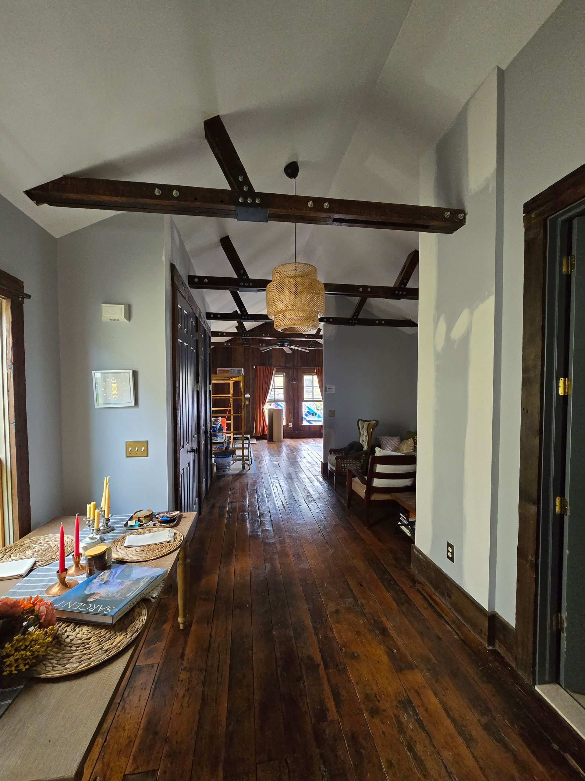 Narrow hallway with wooden floors, exposed beams, and a woven pendant light leading to a brighter room.