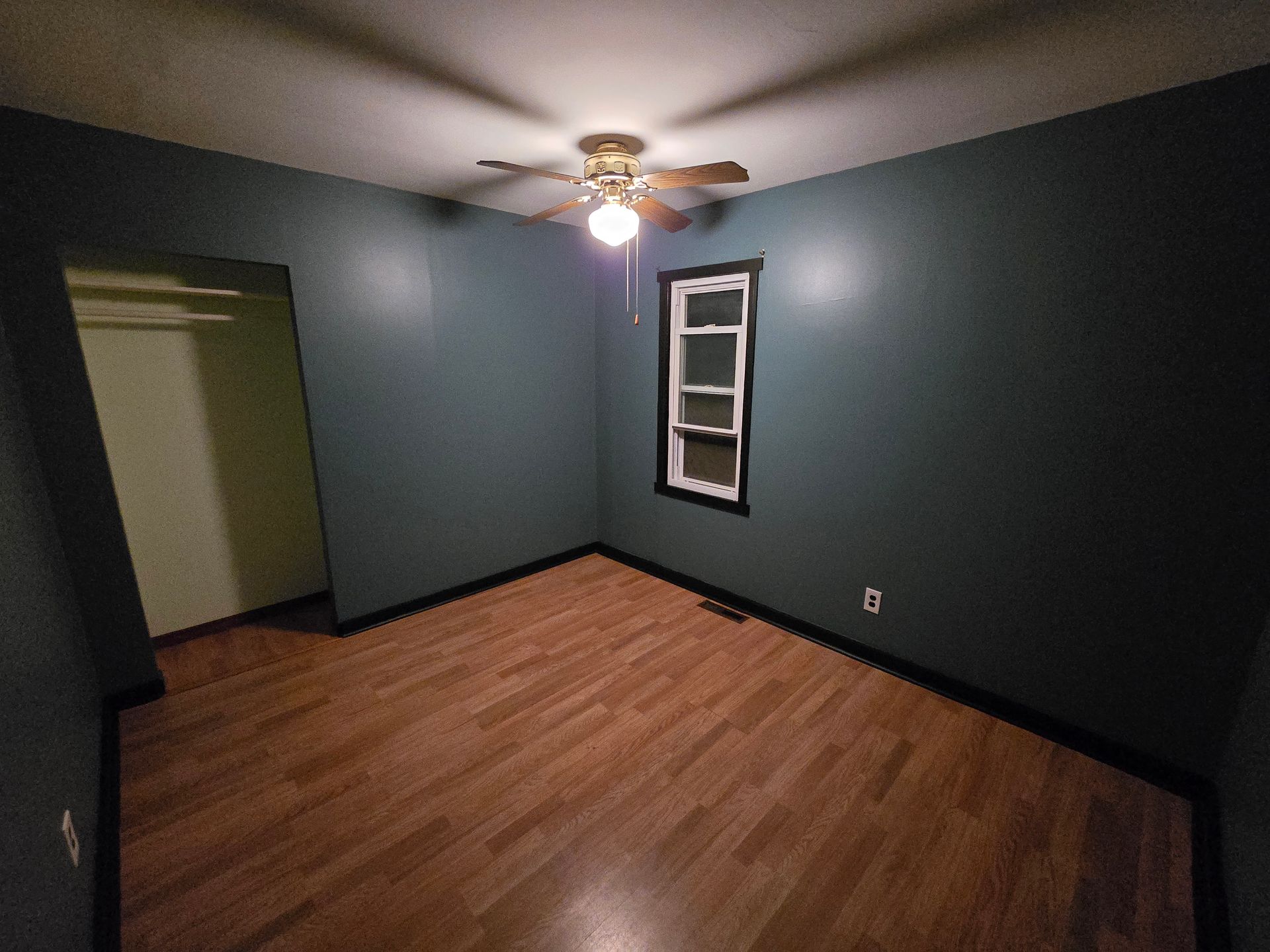 Empty room with dark blue walls, wood floor, ceiling fan light, and a small window.