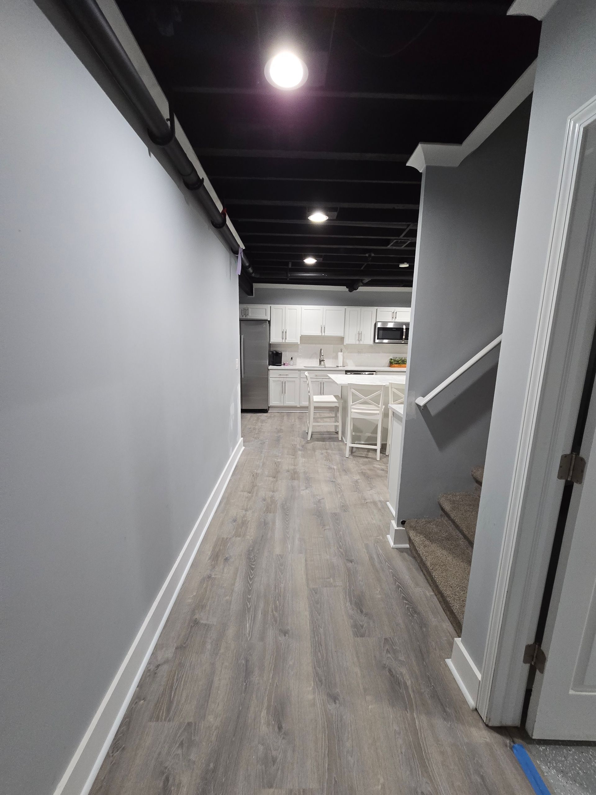Narrow hallway with gray walls and wood floor leading to a bright kitchen area and staircase.