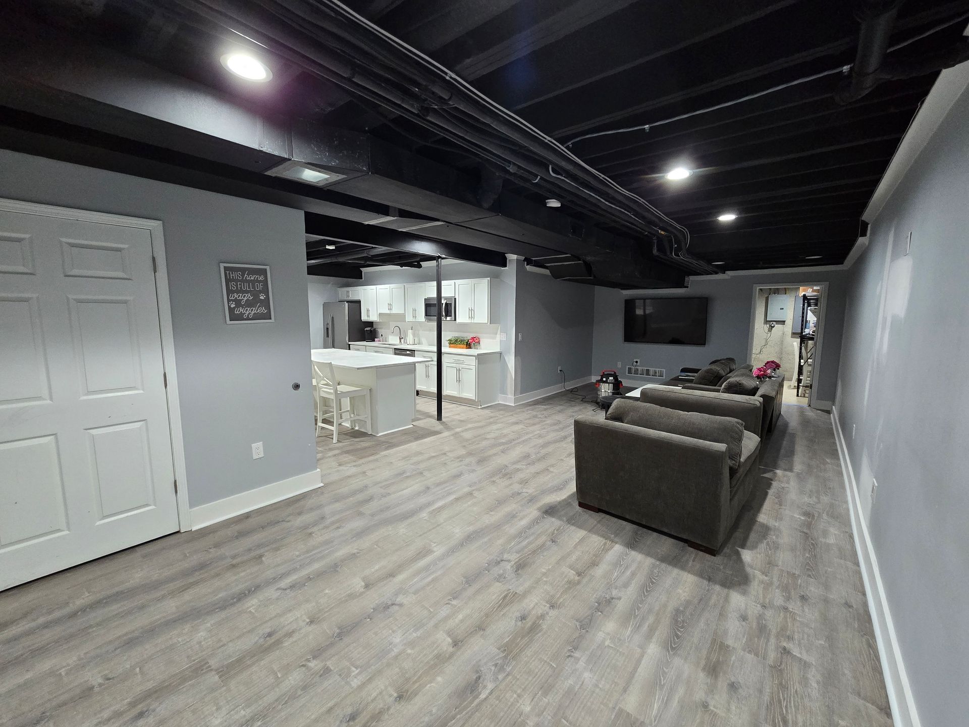 Modern basement living room with gray couch, open kitchen, and black ceiling with recessed lights