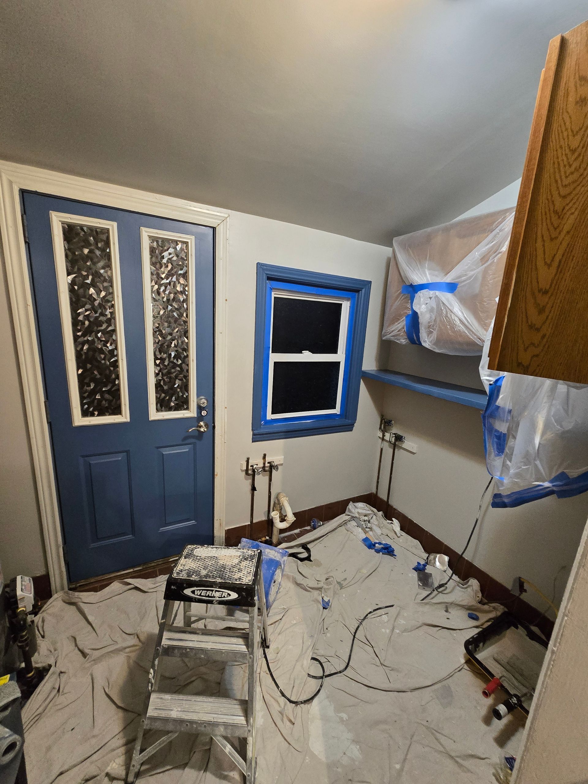Narrow kitchen under renovation with taped windows, blue door, and tools scattered on the floor.