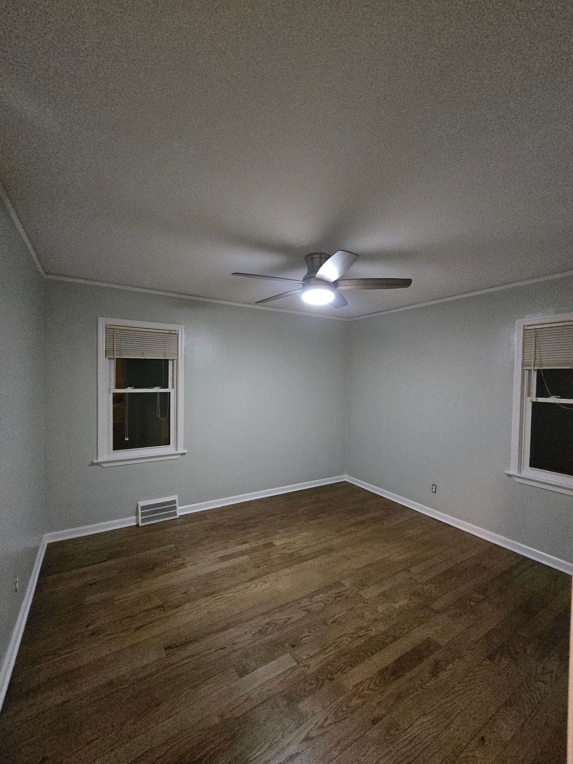Empty white room with wood floors, two windows, and a ceiling fan with lights.
