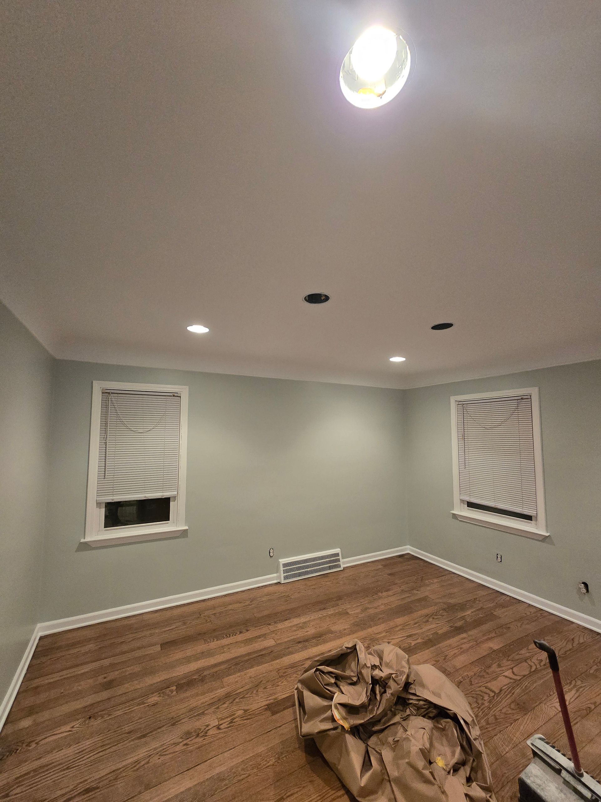 Empty room with gray walls, wood floors, recessed lights, and two windows with white shades.