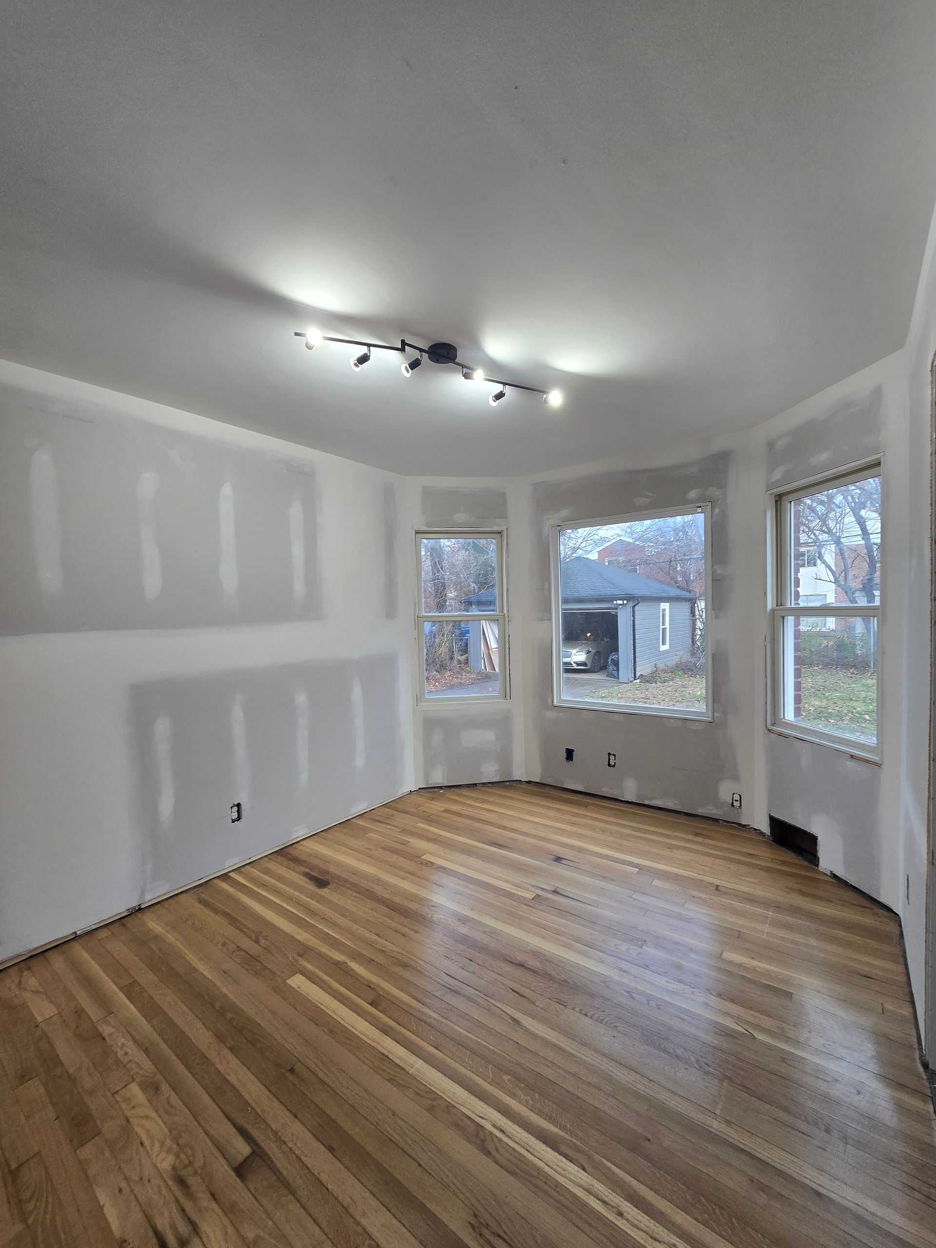Empty room with hardwood floors, unfinished walls, and three windows letting in daylight.