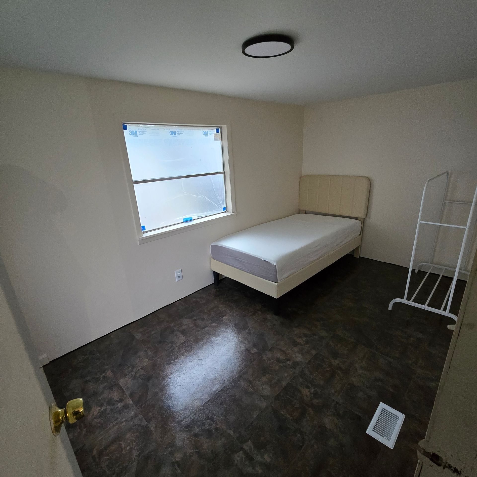 Small empty bedroom with a single bed, window, dark floor, and a white drying rack.
