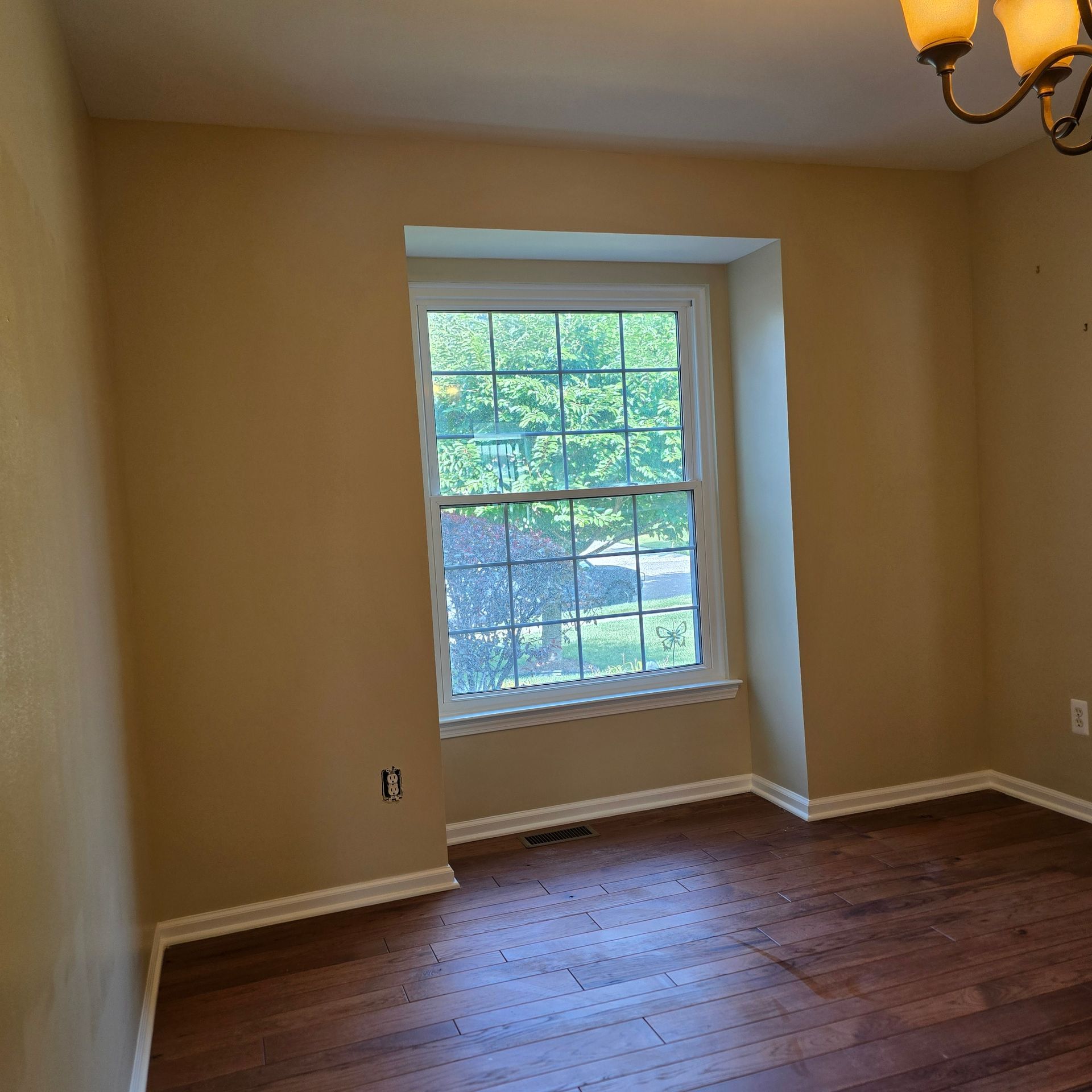 Empty beige room with hardwood floor and a single window overlooking greenery.