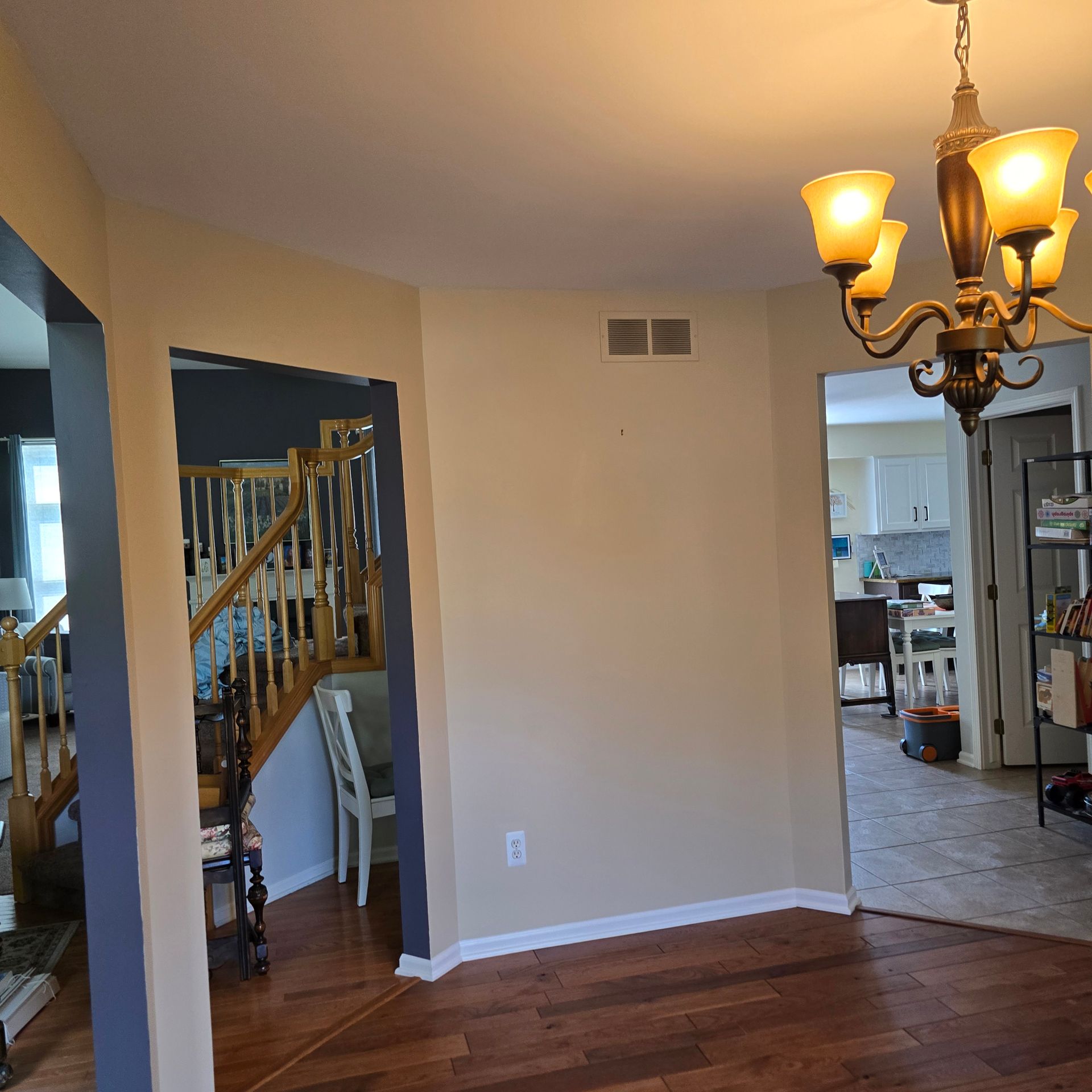 Empty living room with hardwood floors, beige walls, staircase, and chandelier leading to a kitchen area.