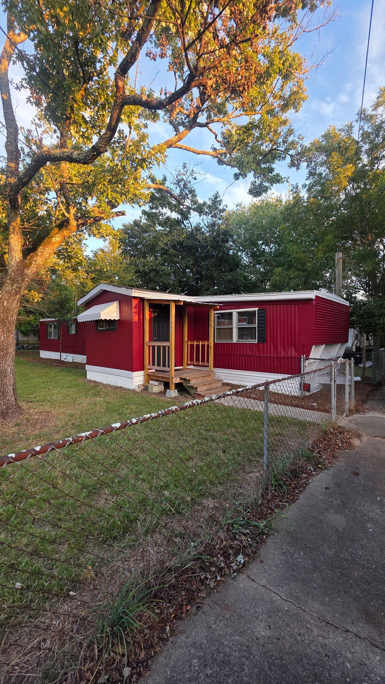 Red Trailer Home with porch in a leafy yard, bordered by trees and a sidewalk at sunset