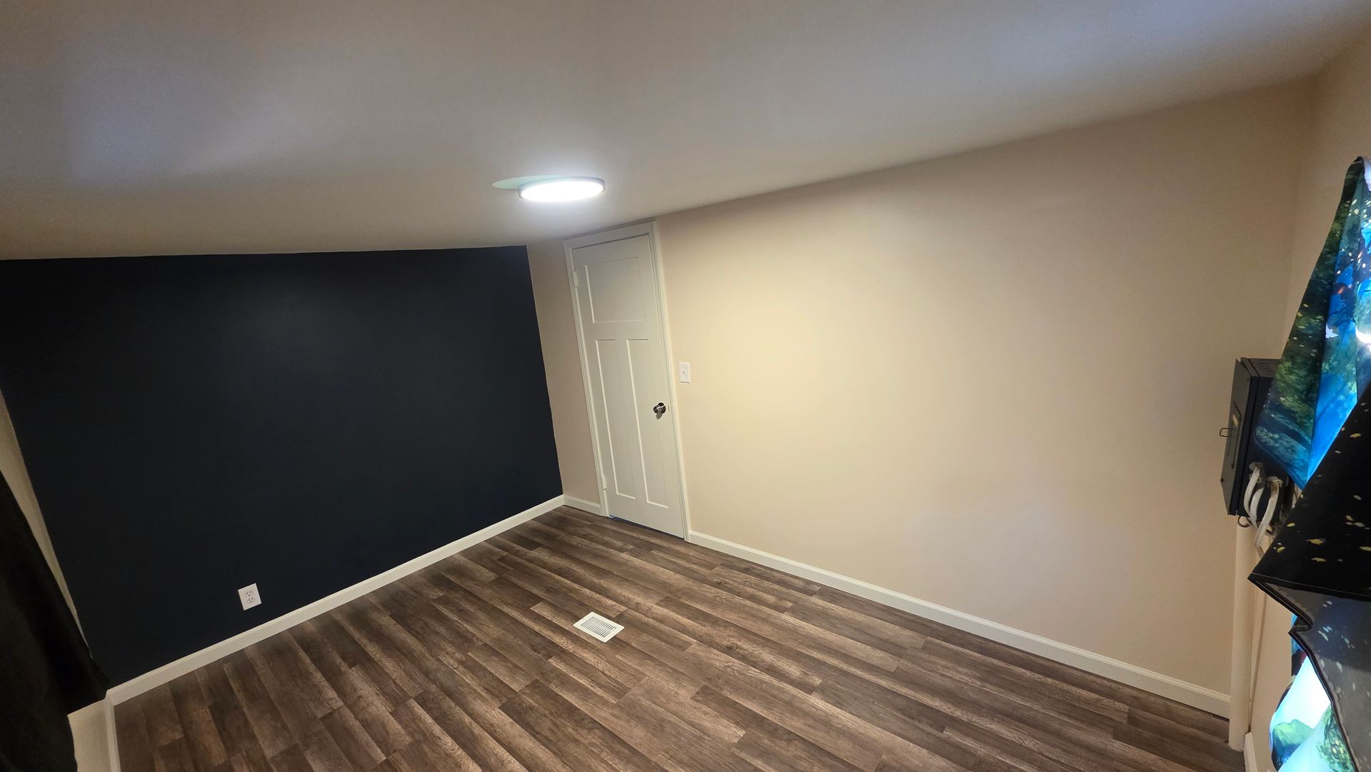 Empty room with dark blue accent wall, beige walls, wood floor, and ceiling light.