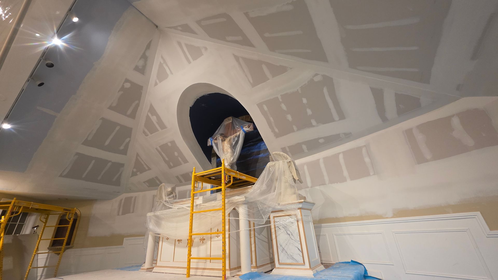 Interior under construction with drywall, scaffolding, and a large circular ceiling opening covered in plastic.