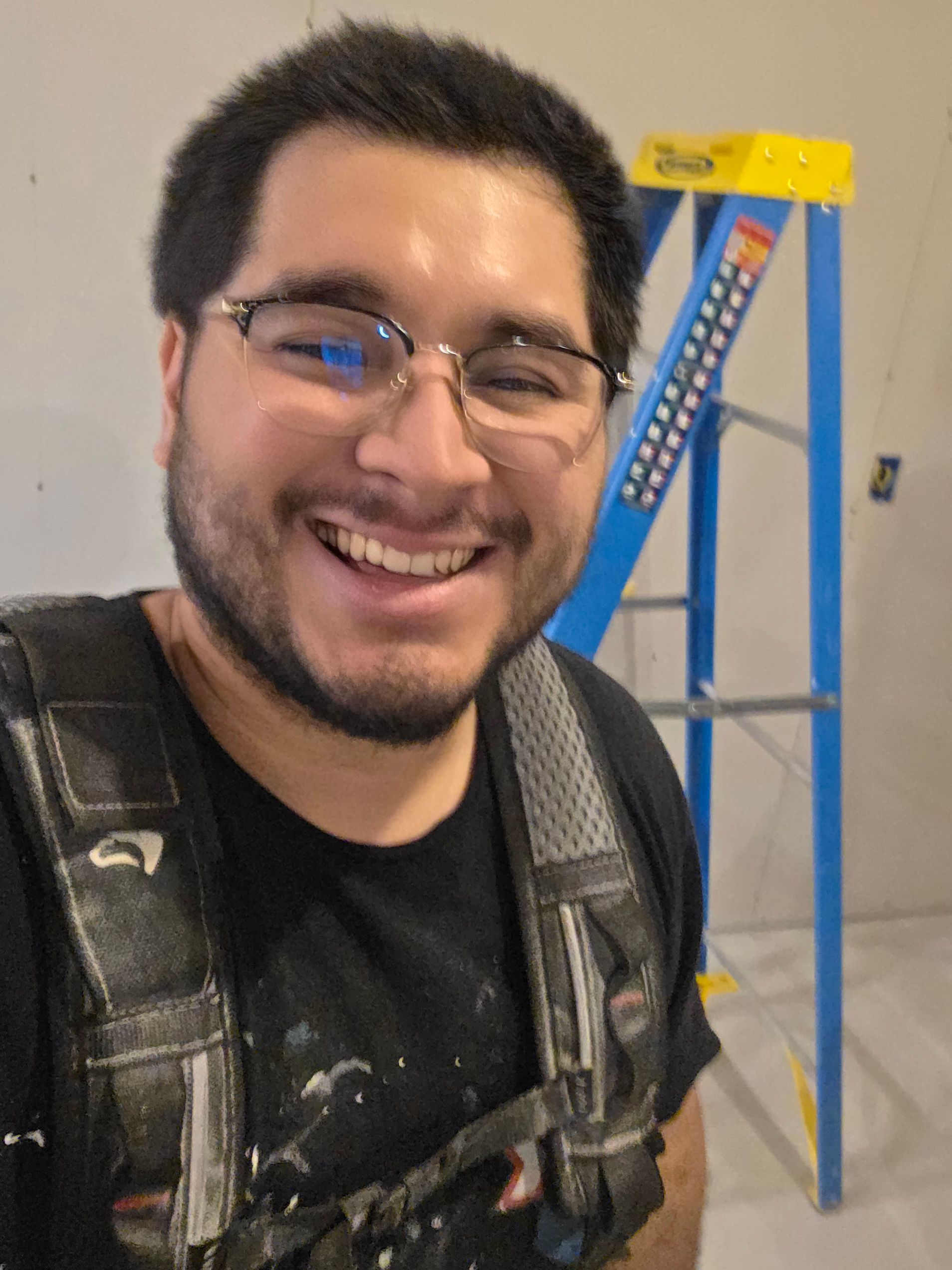 Smiling man in safety harness standing indoors beside a blue ladder on a wall platform
