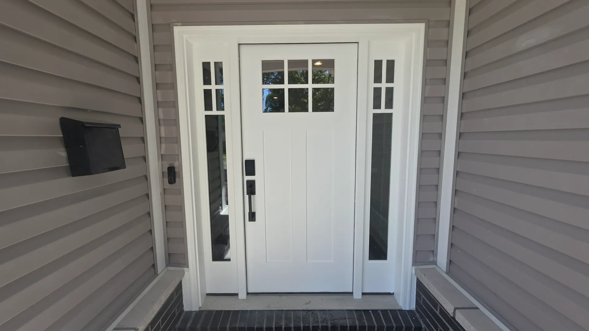 White front door with sidelights and a small window, centered on a covered porch with gray siding and black floor mat.