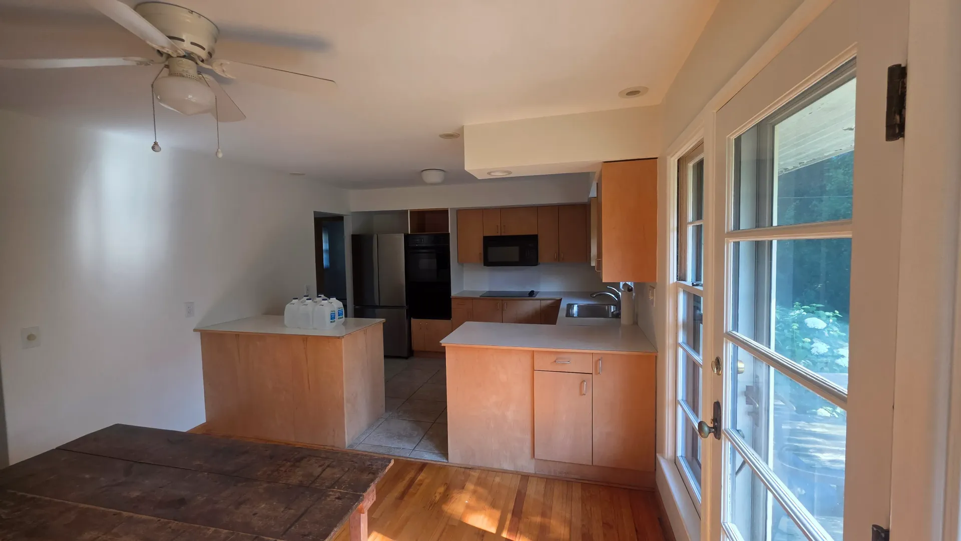 Open kitchen and living room with wood cabinets, ceiling fan, and sunlight through large windows