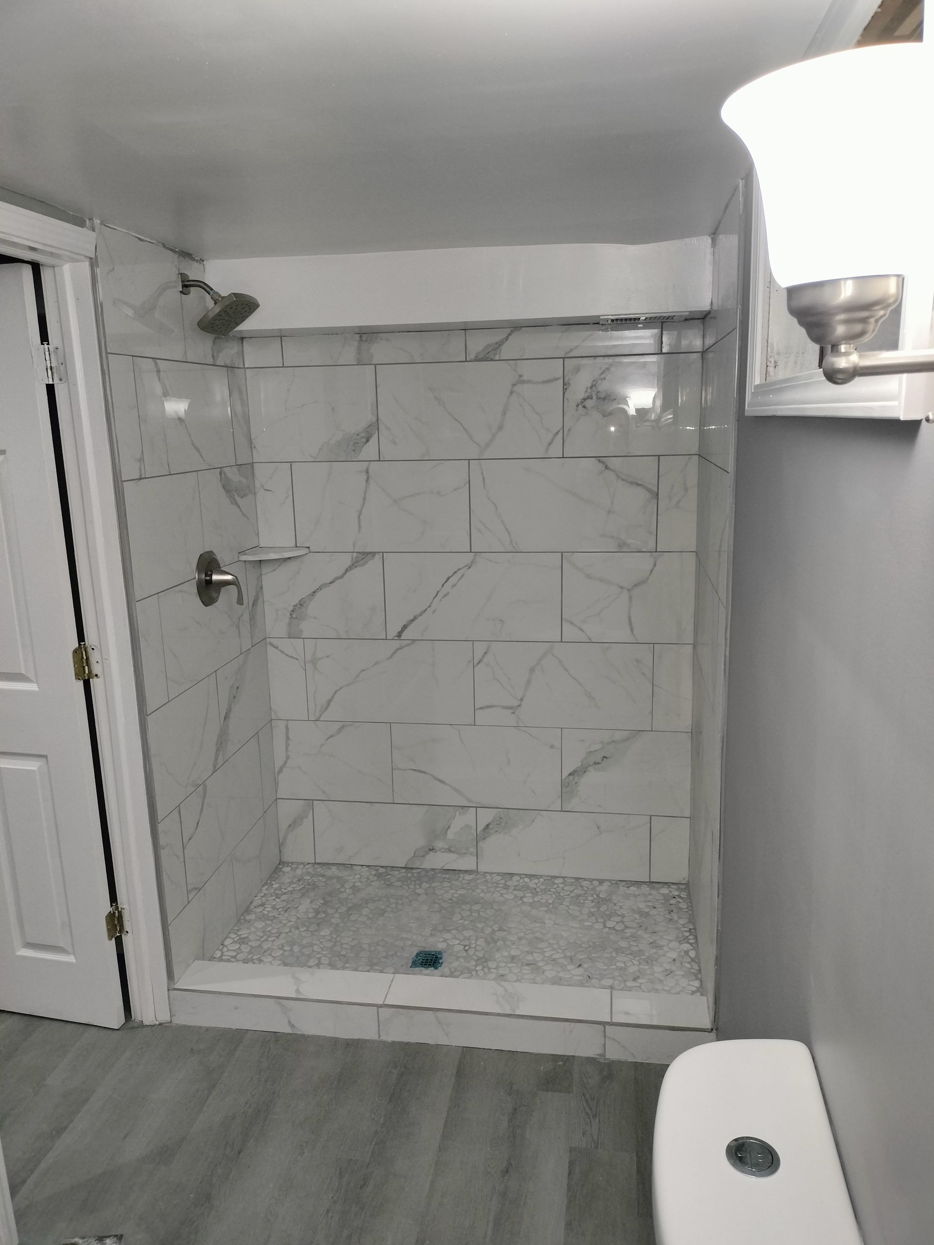 Small bathroom shower alcove with white tile walls, gray floor, and a toilet edge in the foreground