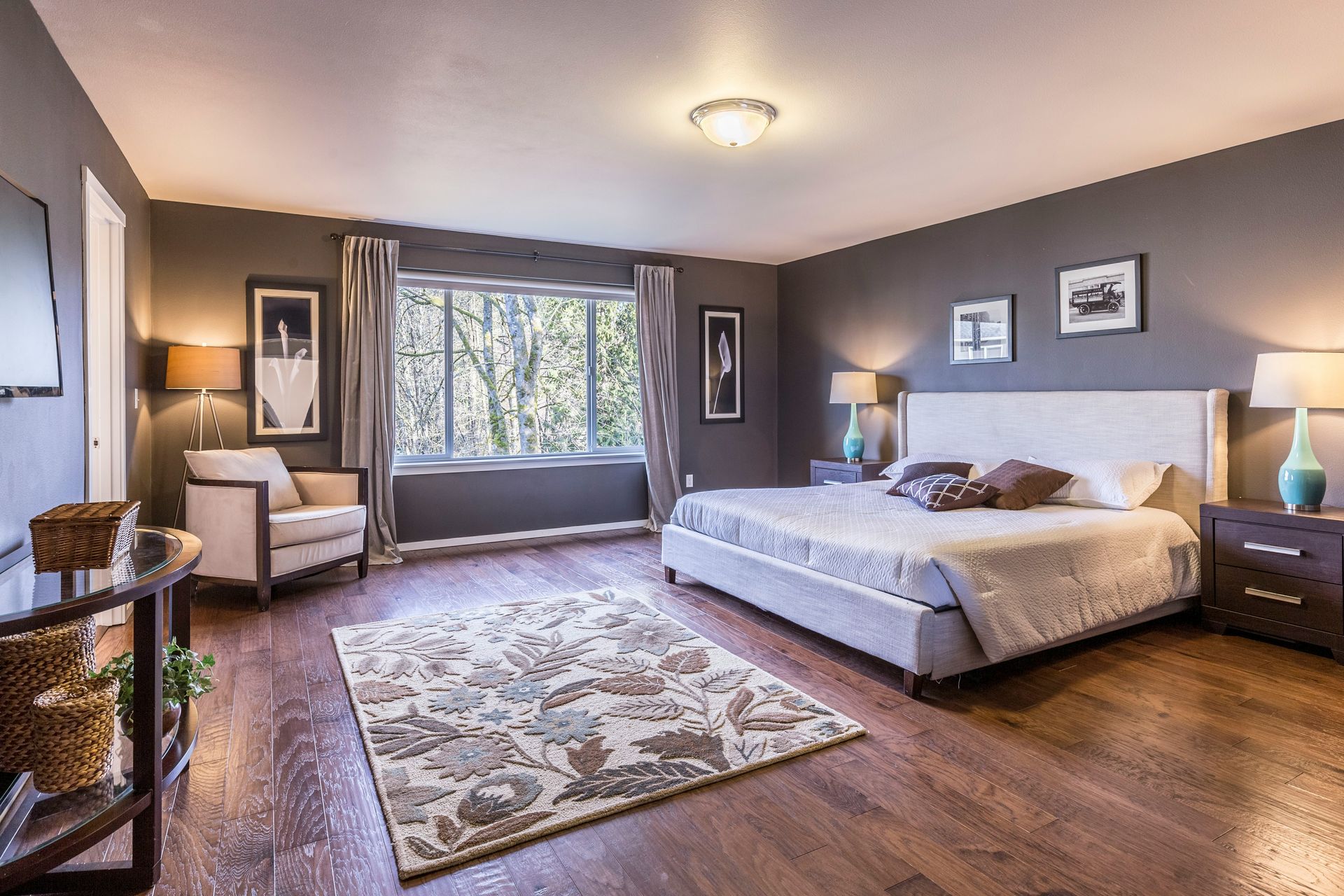 Spacious bedroom with a large bed, armchair, patterned rug, and window seat in soft gray tones.