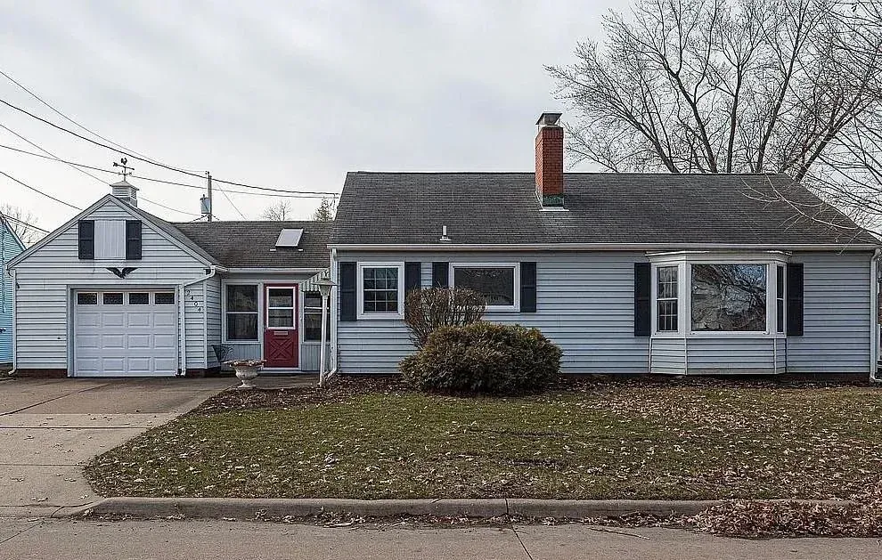 Small gray house with a red front door, chimney, garage, and leafless trees in front
