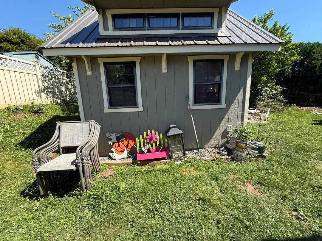 Small gray garden shed with two windows, wicker chairs, and potted flowers in a grassy yard.