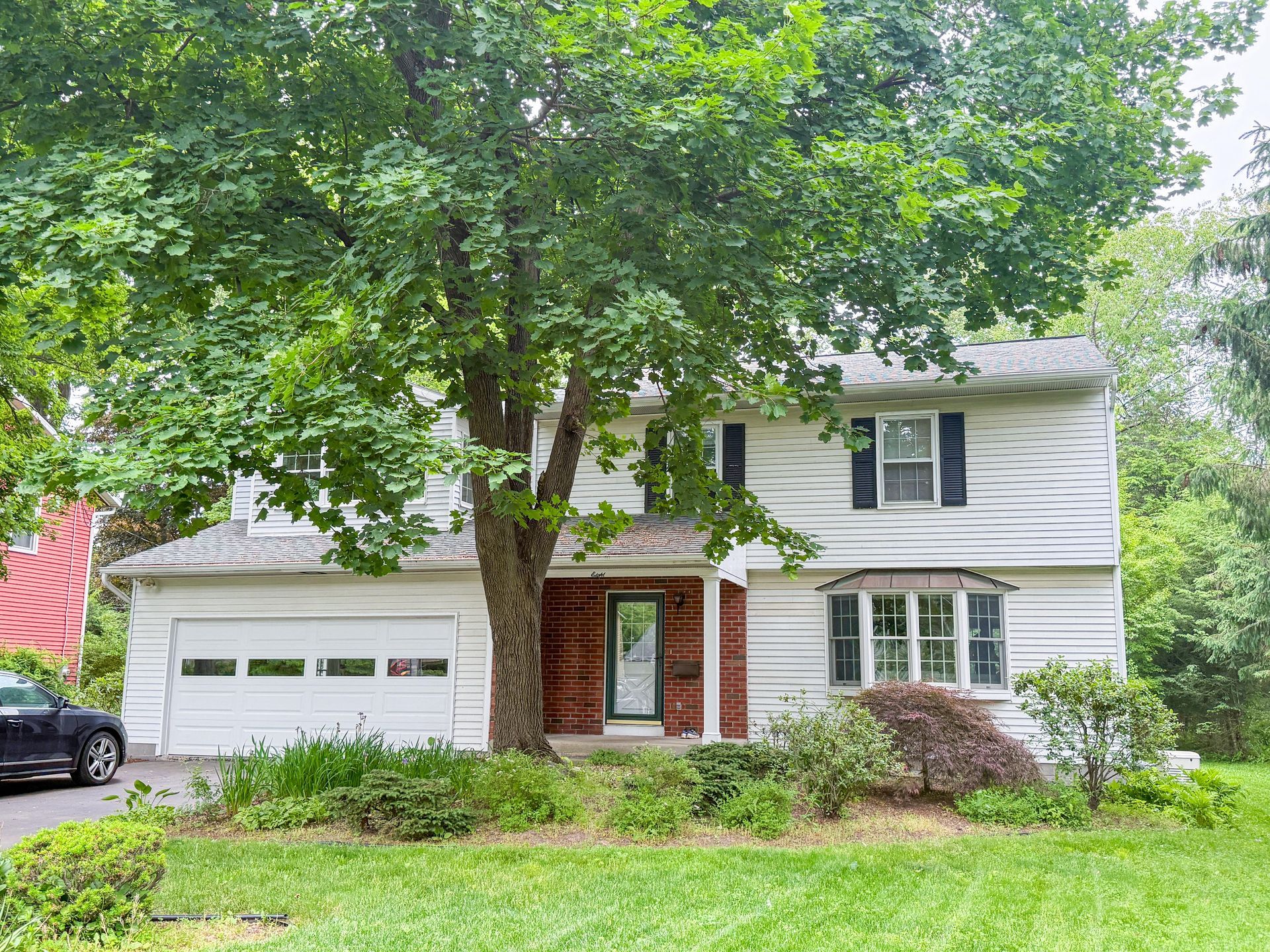 Two-story white house with a garage, large tree, and front lawn surrounded by greenery
