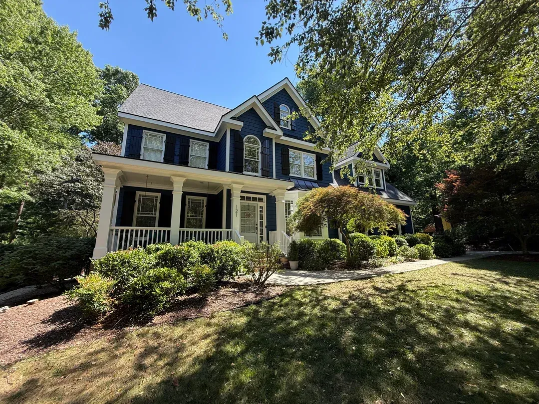 Two-story blue house with white porch, surrounded by trees and a sunlit front yard