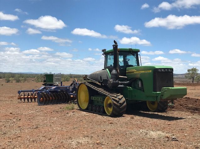 A green John Deere tractor with rubber tracks pulls a dark disc plow across a dry, brown field under a cloudy blue sky.