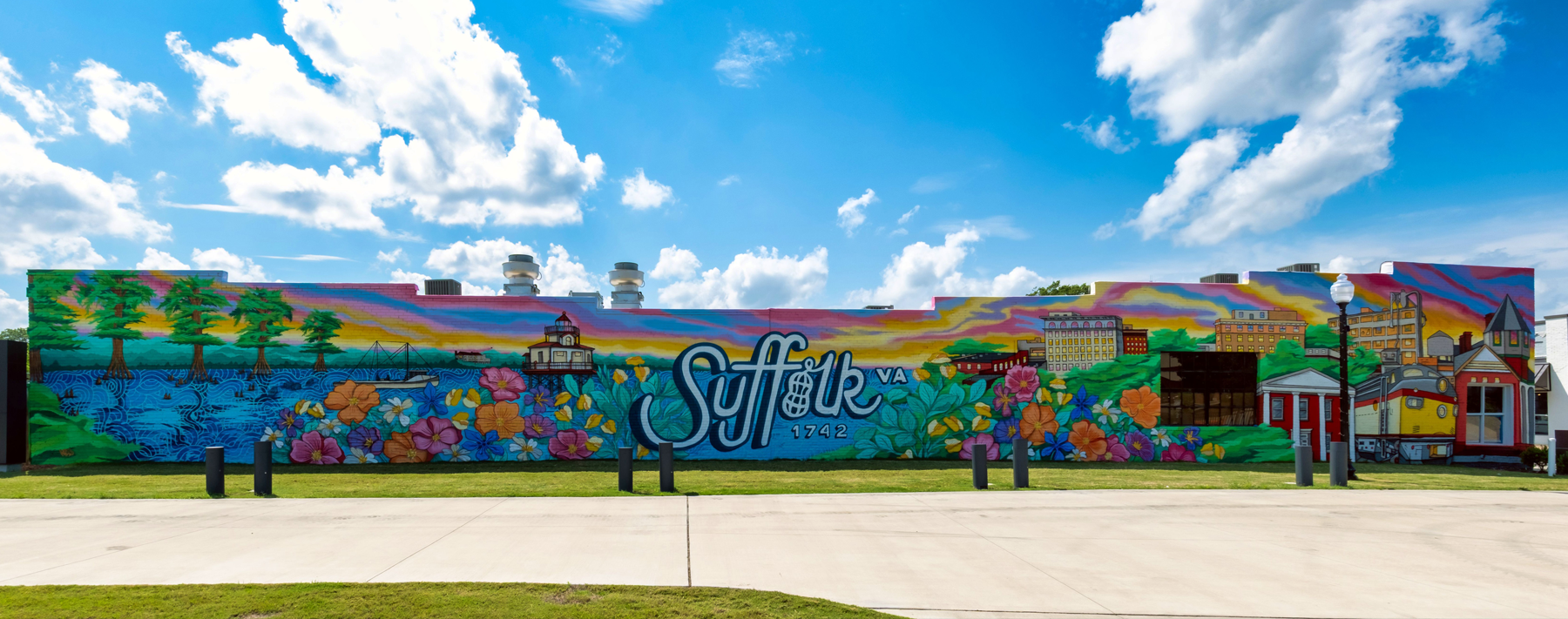 Colorful mural on a long building spelling “Suffolk, VA” with a blue sky and grassy foreground