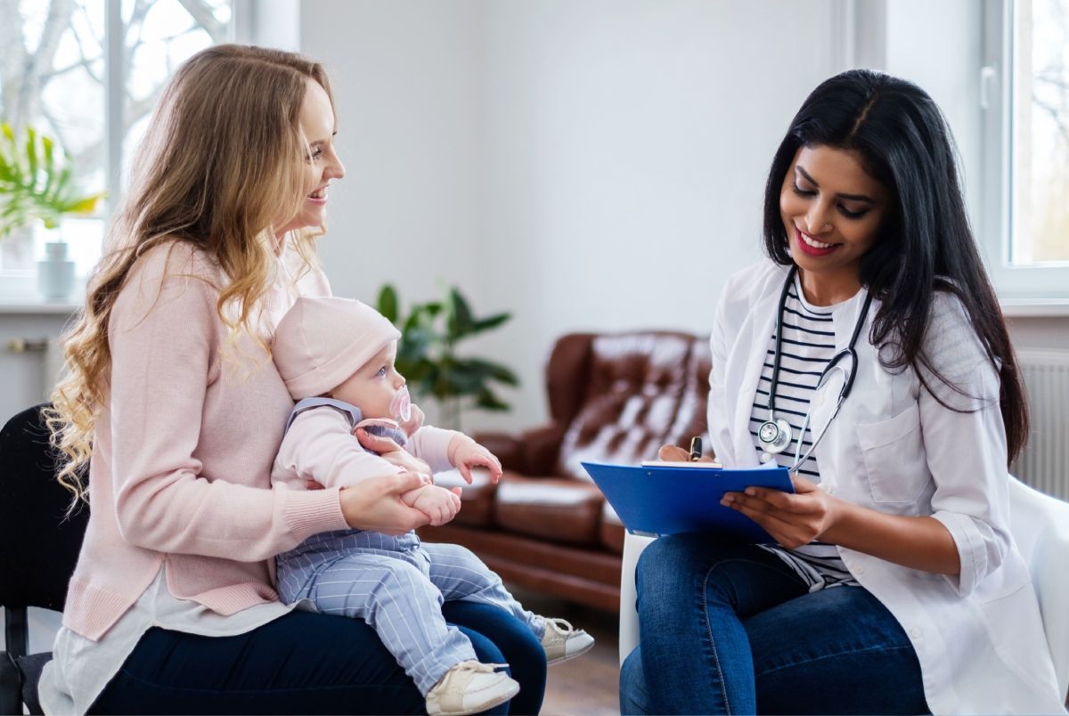 Woman holding baby consults with a doctor. The doctor smiles while holding a clipboard. In an office setting.