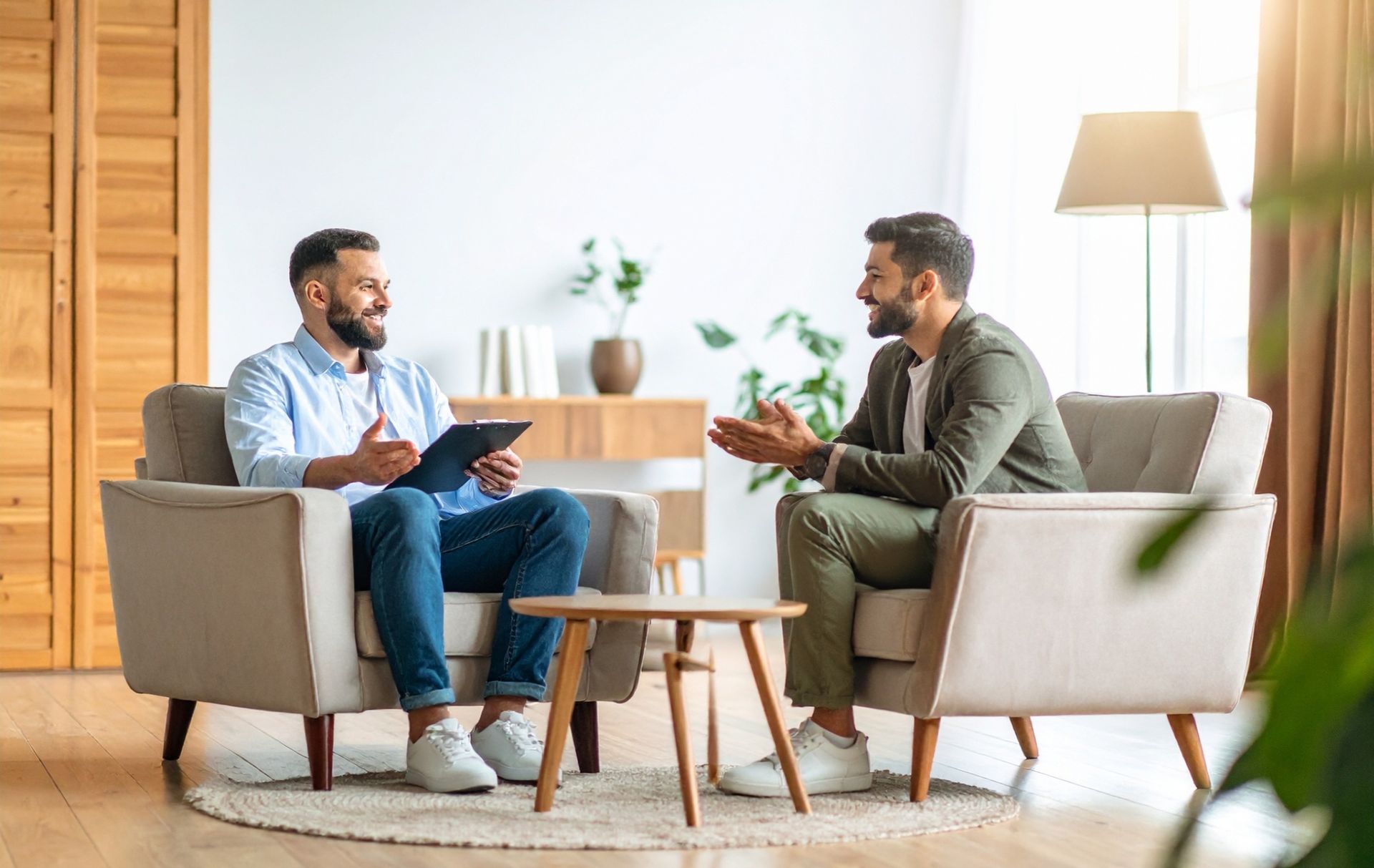 Two men talking in a bright living room, seated in armchairs with a small table between them.