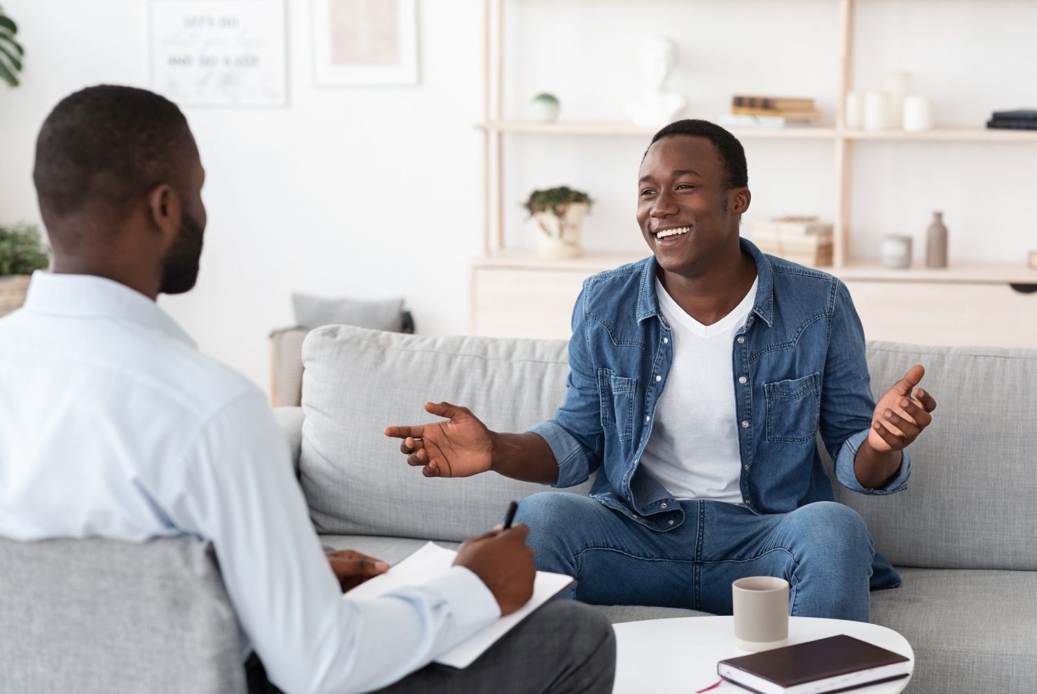 Two people talking on a gray couch in a bright living room, one smiling and gesturing with hands