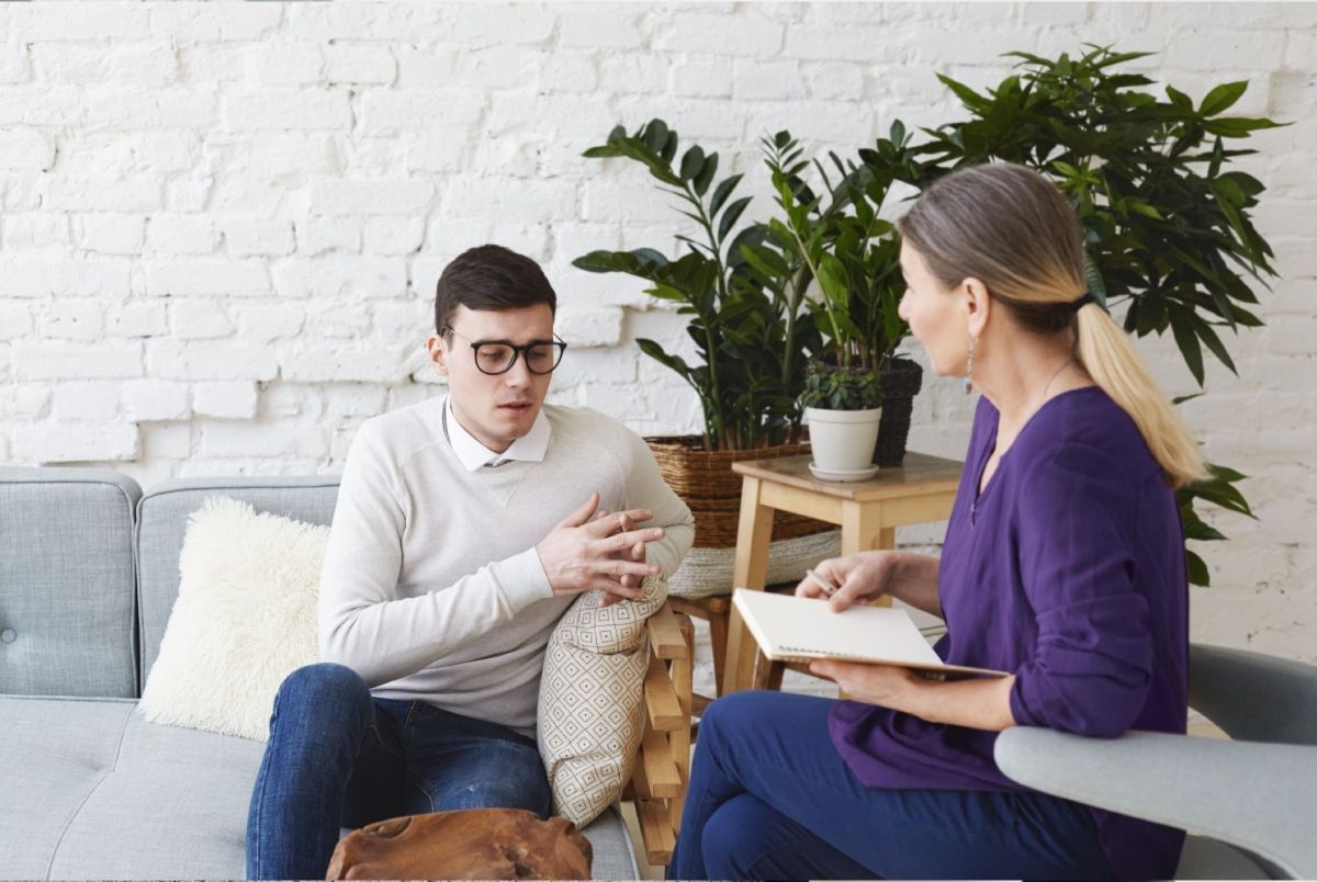Two people talking in a bright living room, one holding a notepad on a couch.