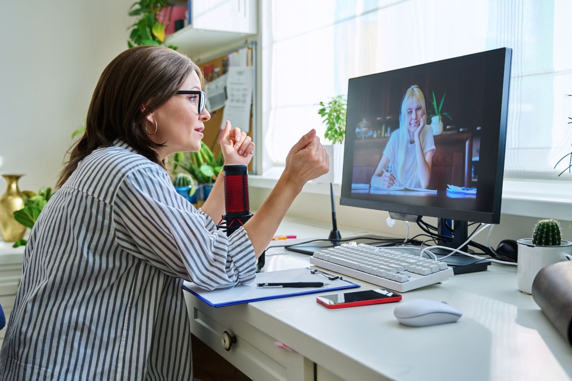 Woman in glasses on a video call, gesturing with hands, sitting at a desk with a computer and papers.