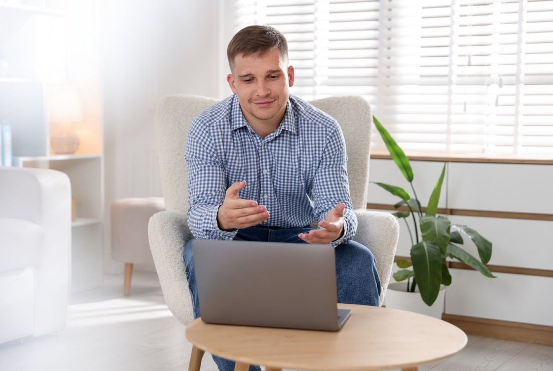 Man in blue plaid shirt sits in chair, using laptop, gesturing, indoors.