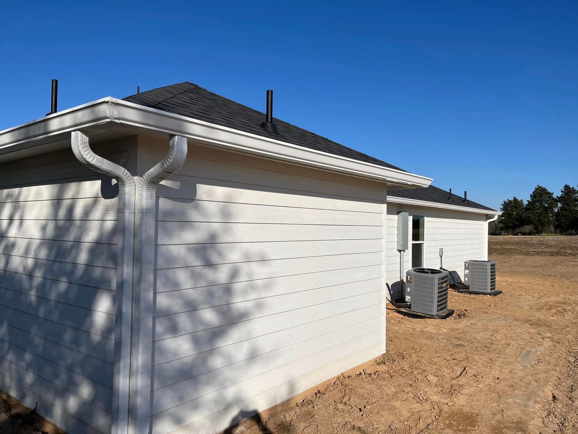 White house exterior with dark roof, gutters, and HVAC units beside a dirt yard under a blue sky