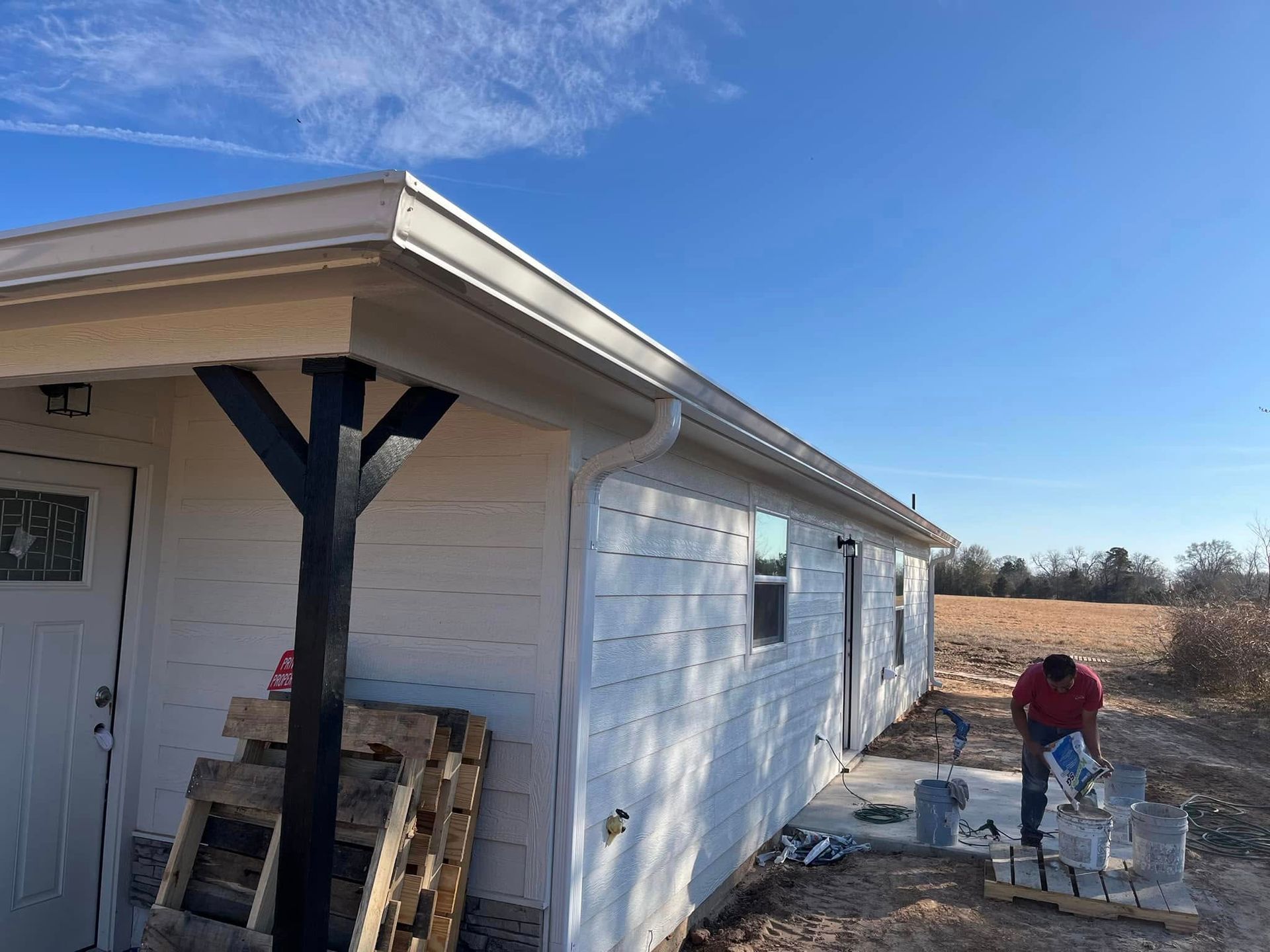 Person painting the side of a white house beside a garage in a rural yard