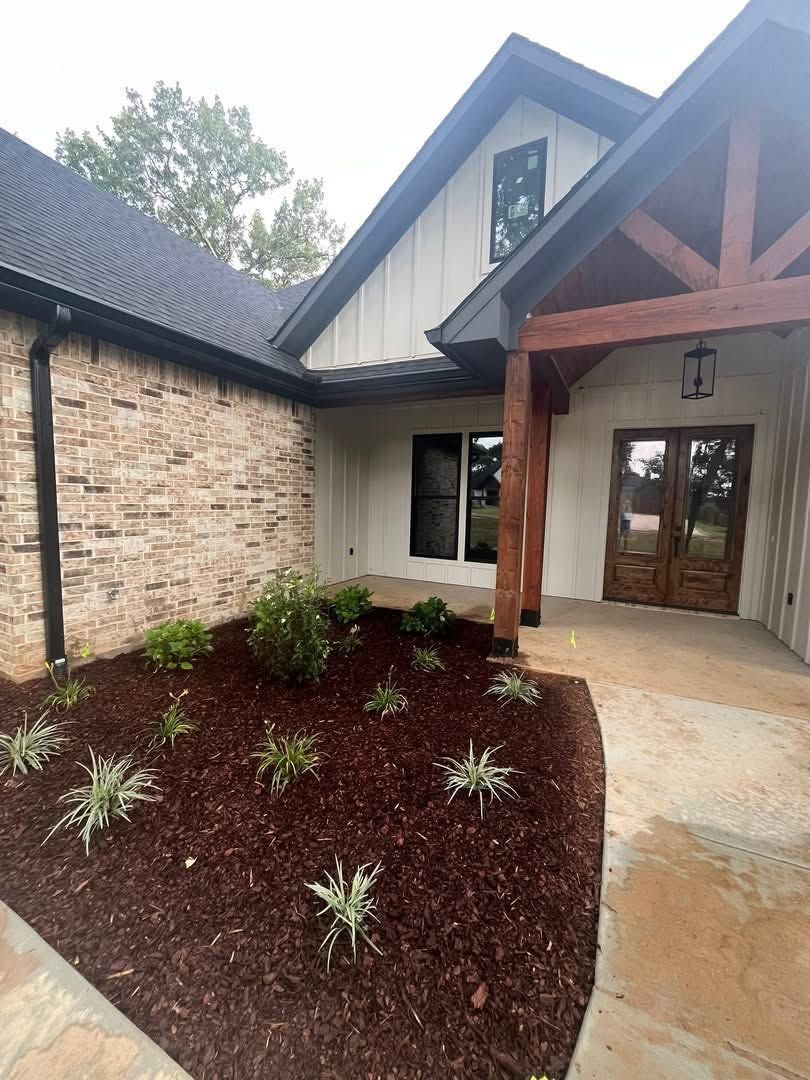 Stone house entrance with covered porch, wood beams, and a landscaped mulched garden bed with small plants.