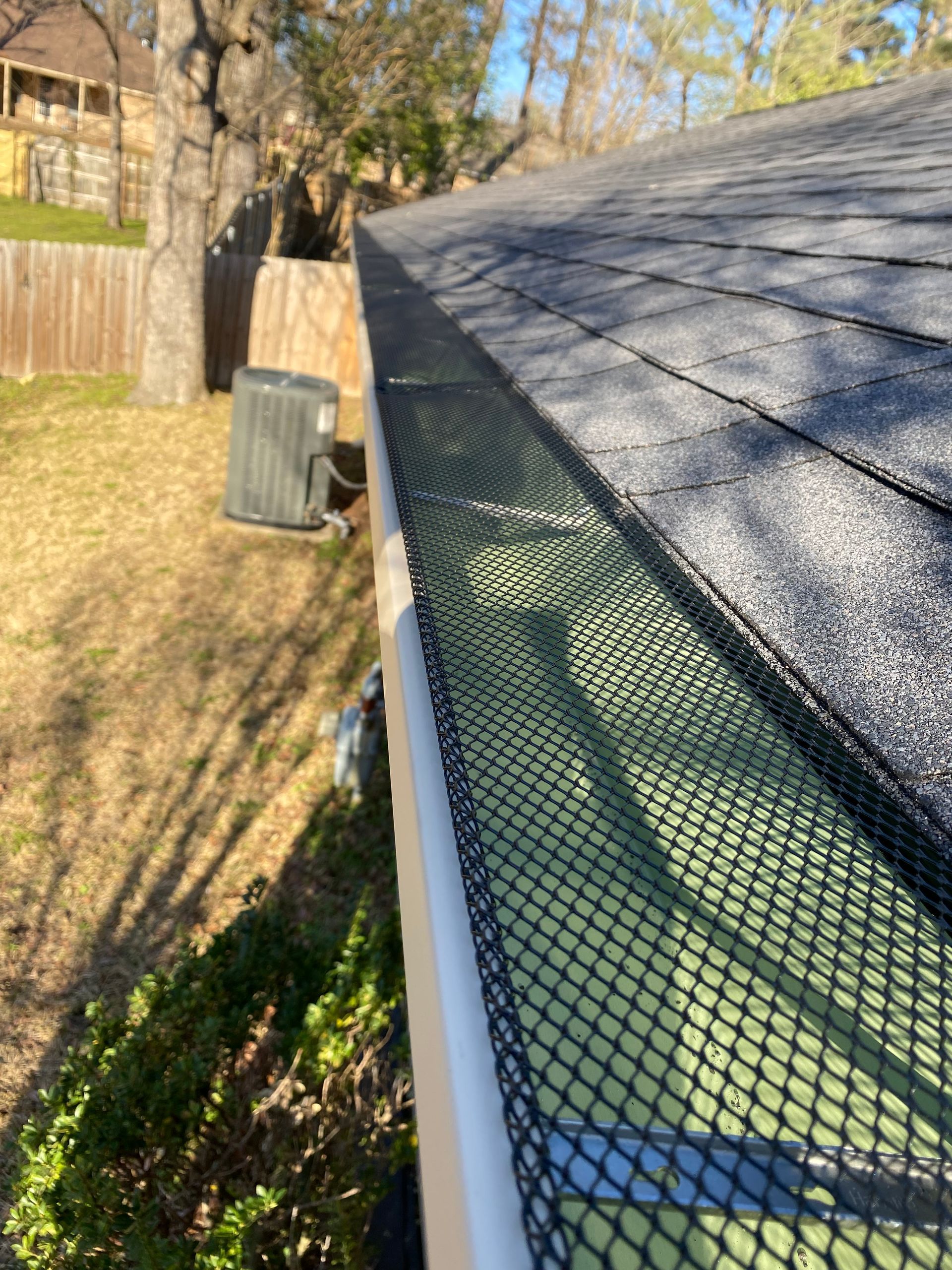Gutter with leaf guard along a shingled roof edge beside a backyard fence and trees
