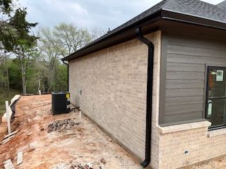 Side of a brick building with downspout, muddy ground, and surrounding trees under a cloudy sky