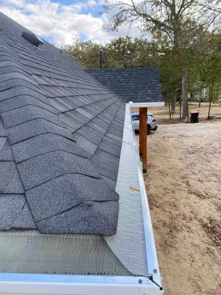 Roof gutter beside gray shingle roof, viewed along the eave in a sandy yard.