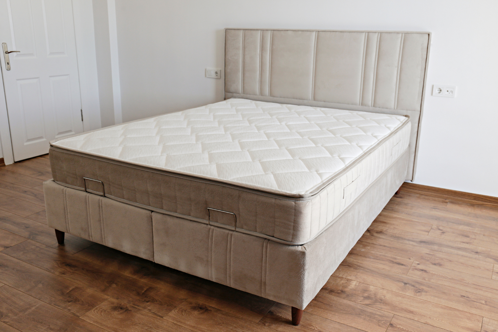 Beige upholstered bed with tufted headboard and mattress in a white bedroom with wood flooring