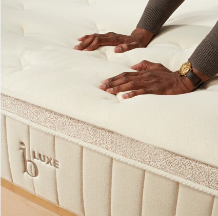 Beige upholstered bed with tufted headboard and mattress in a white bedroom with wood flooring