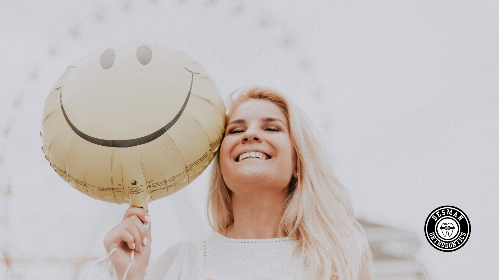 Smiling blonde woman holding a smiley-face balloon against a light background