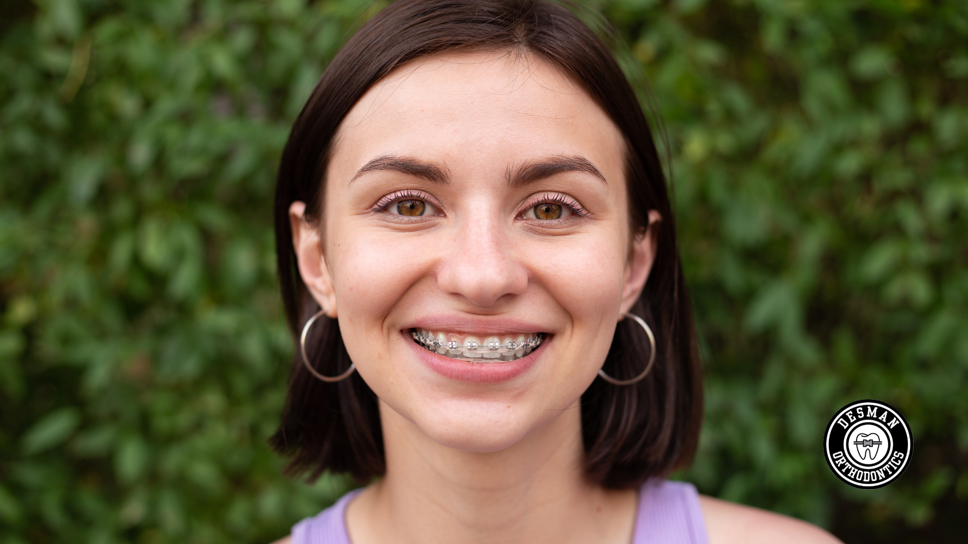 Smiling person with braces and hoop earrings in front of green foliage