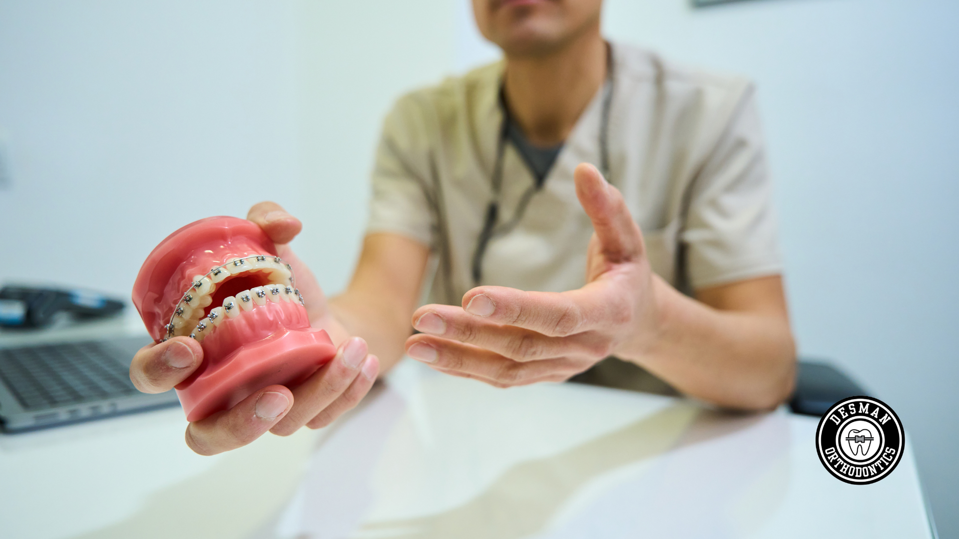 Dentist showing a pink dental model to a patient in a clinic, with laptop and white desk in the background