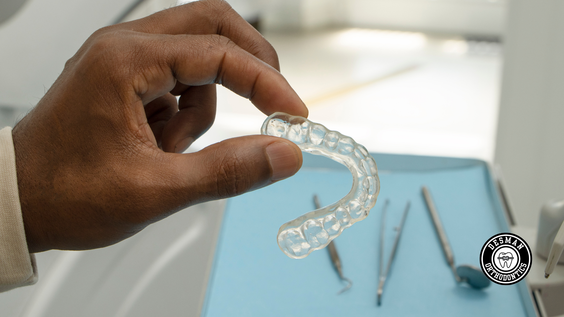 A hand holding a clear dental aligner over a blue tray with dental tools nearby.