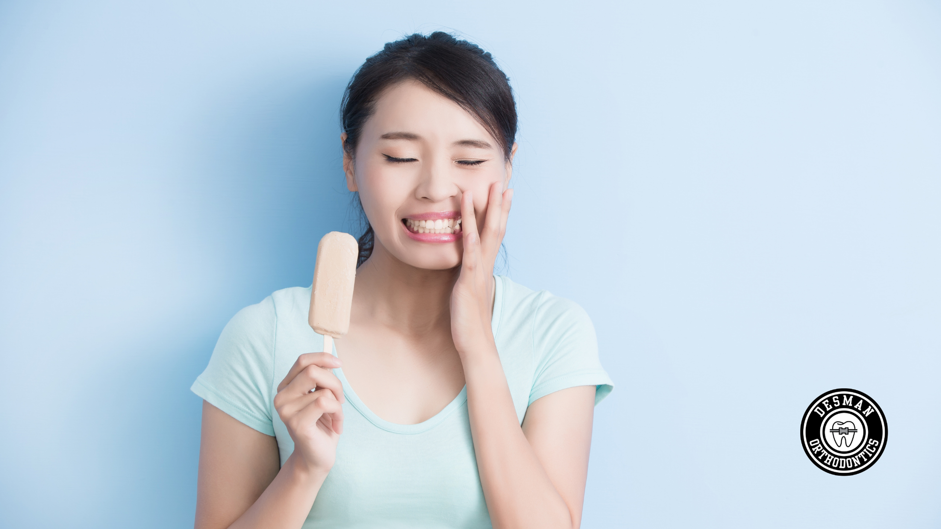 Woman touching cheek while holding a popsicle, wincing against a blue background